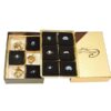 The open box of Aqua Marine Crystal Bracelets with a beige lid displays nine compartments; some contain gold-wrapped chocolates, while others hold shiny chocolates.