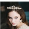 A woman wearing an ivory tiara accented with champagne and gold pearls and opals, complemented by matching earrings, looks down with a neutral expression.